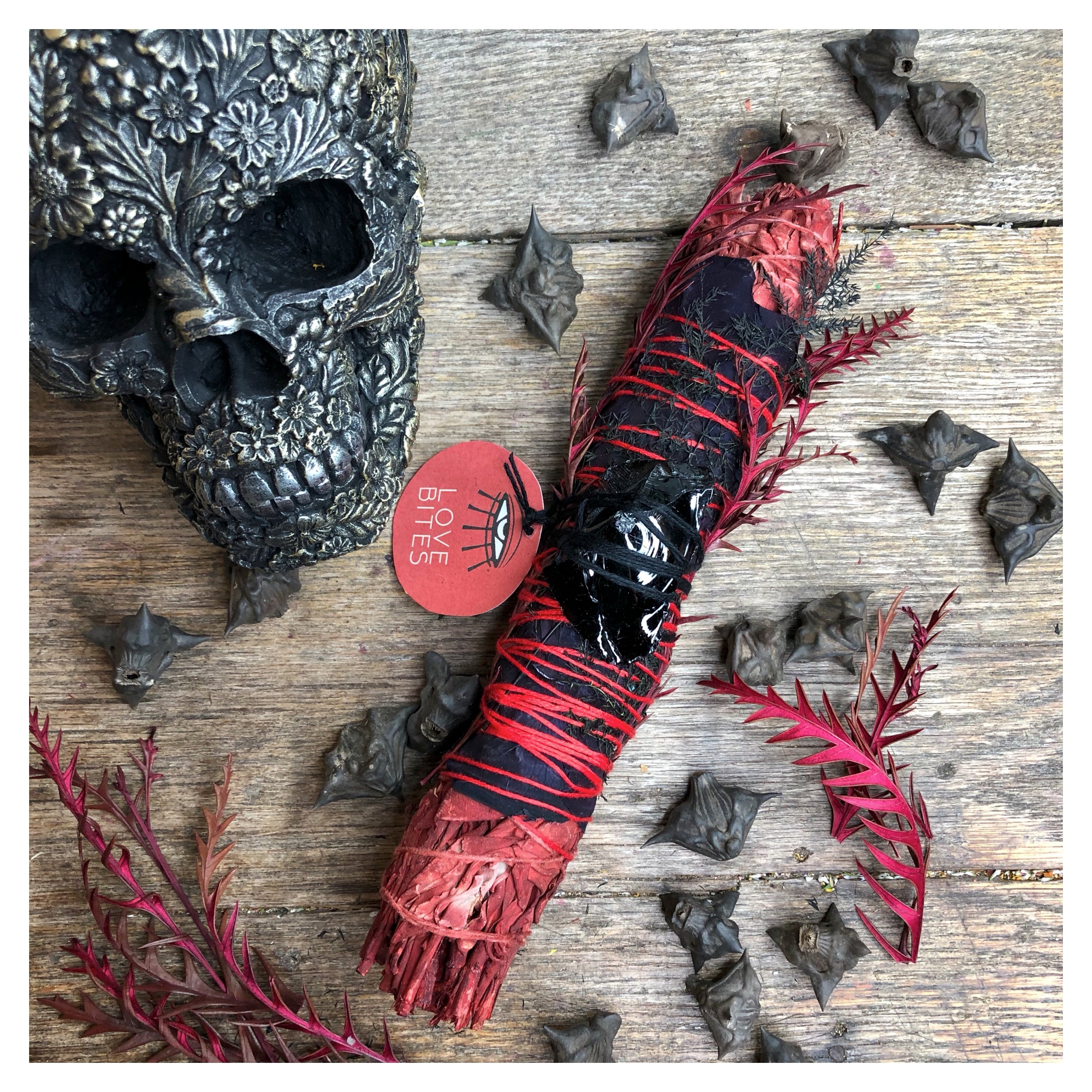 large 9" dragon's blood sage smoke cleansing smudge bundle with black rose petals, spike red dried florals and a large obsidian crystal attached with skull and bat wing nuts in background on wood surface