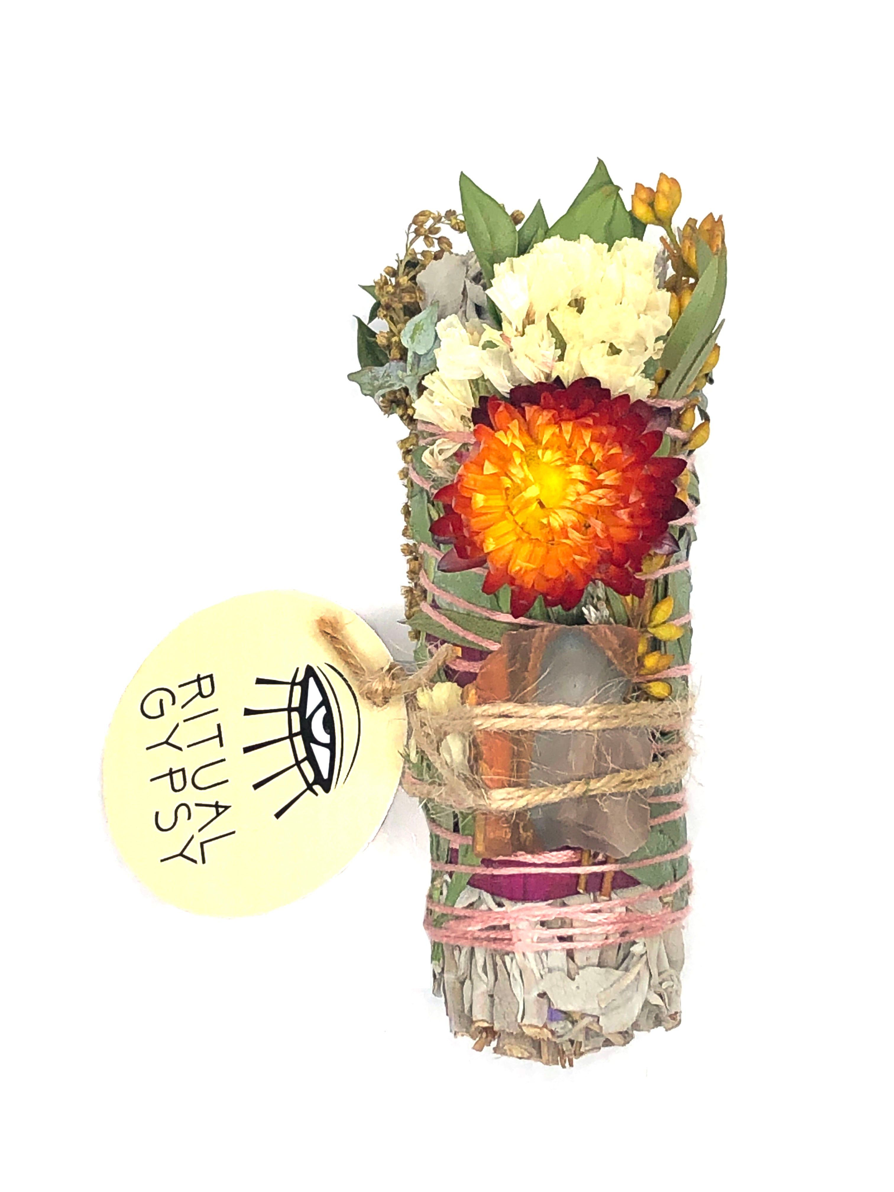 white sage smoke cleansing smudge bundle wrapped in florals with orange red strawflower and orange brown gray desert jasper crystal and round ritual gypsy product tag attached on white background