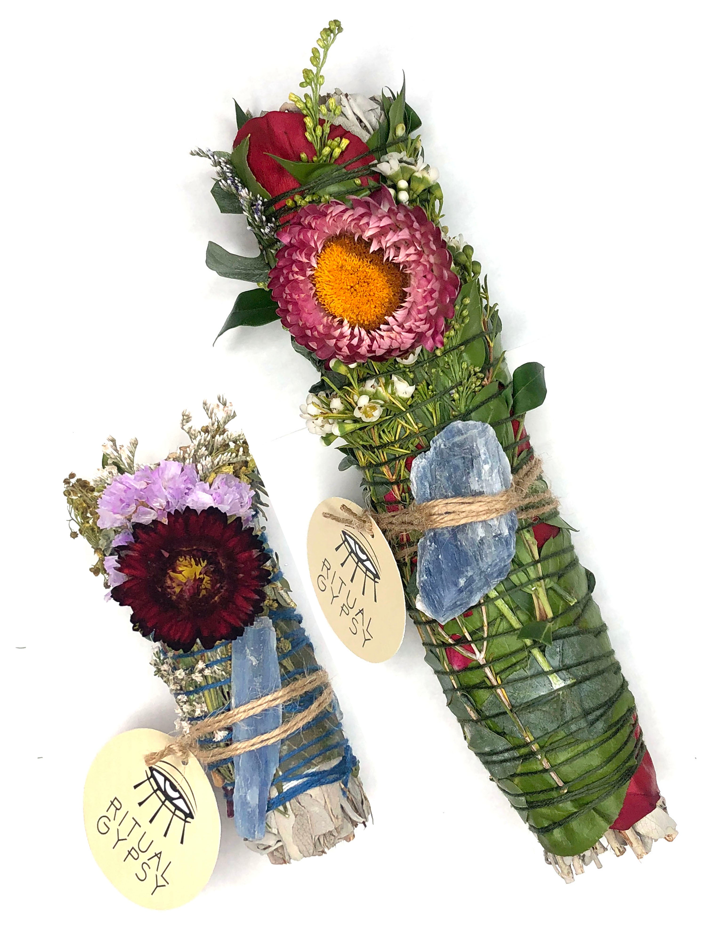 white sage smoke cleansing smudge bundle wrapped in florals with red strawflower and blue kyanite blade crystal and round ritual gypsy product tag attached on white background