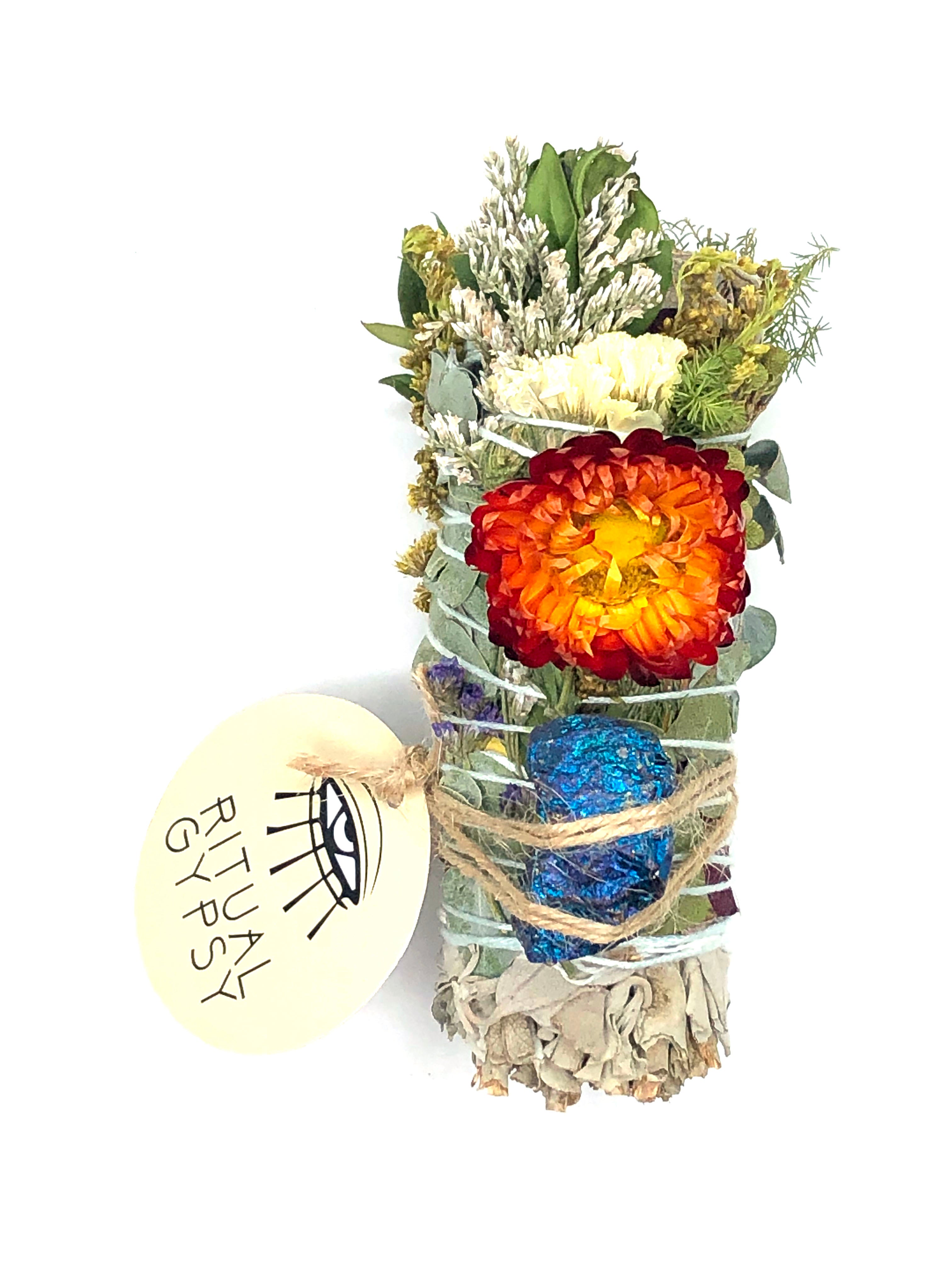 white sage smoke cleansing smudge bundle wrapped in red and yellow rose petals, strawflowers and florals with blue purple peacock ore crystal and round ritual gypsy product tag on white background