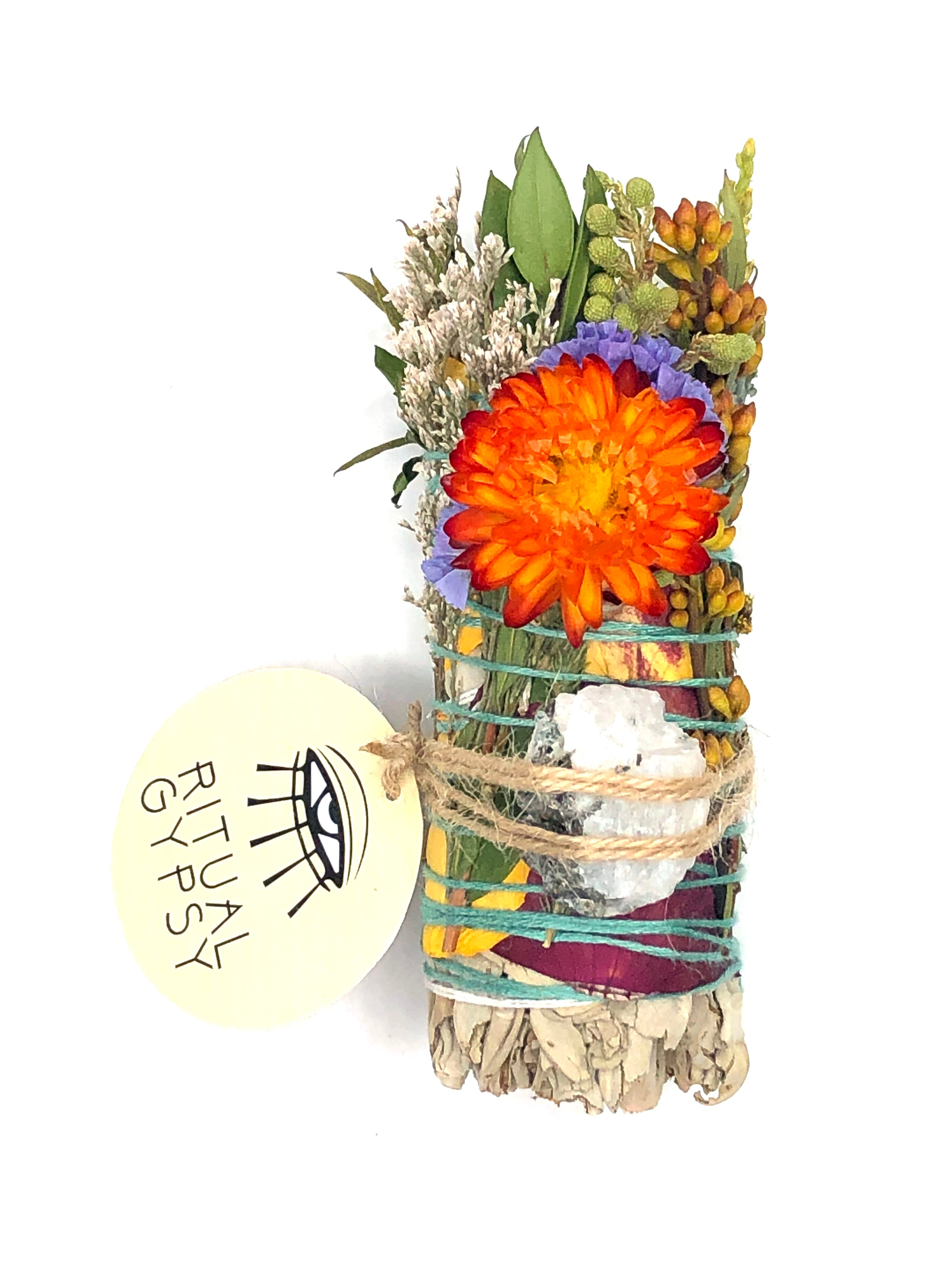 white sage smoke cleansing smudge bundle wrapped in florals with red strawflower and white and black rainbow moonstone crystal and round ritual gypsy product tag attached on white background