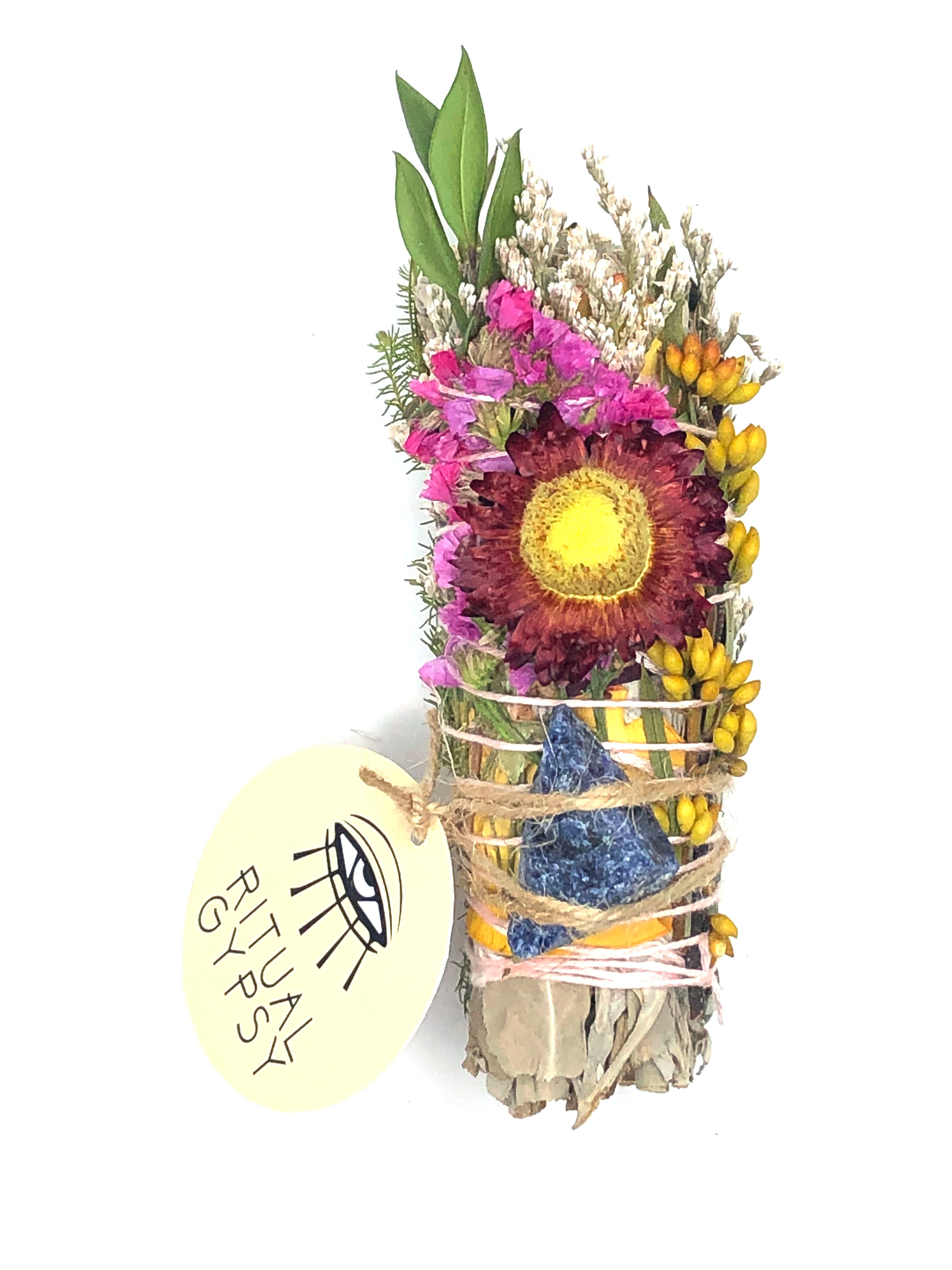 white sage smoke cleansing smudge bundle wrapped in florals with red strawflower and blue sodalite crystal and round ritual gypsy product tag attached on white background