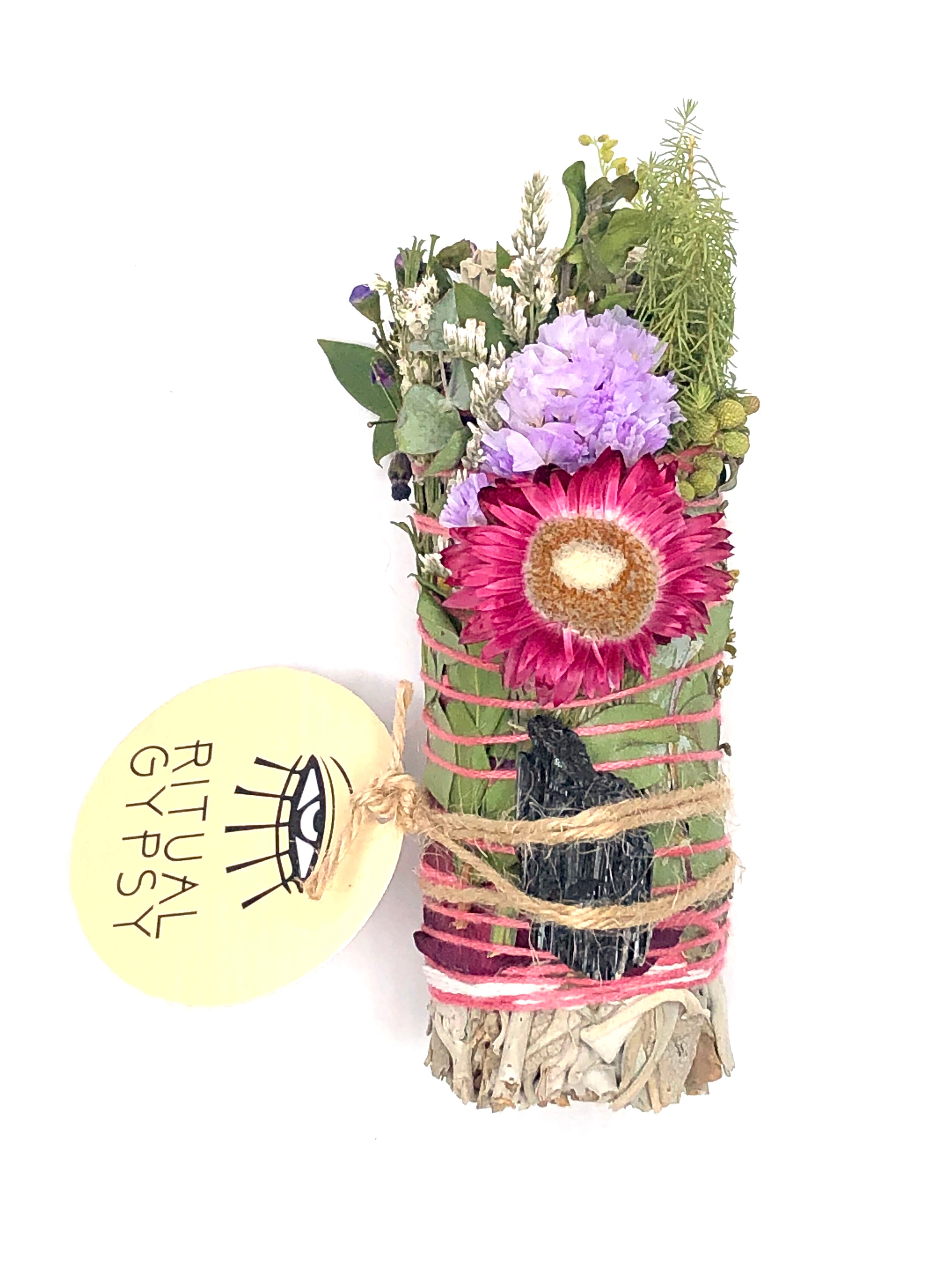white sage smoke cleansing smudge bundle wrapped in florals with orange red strawflower and black tourmaline crystal and round ritual gypsy product tag attached on white background