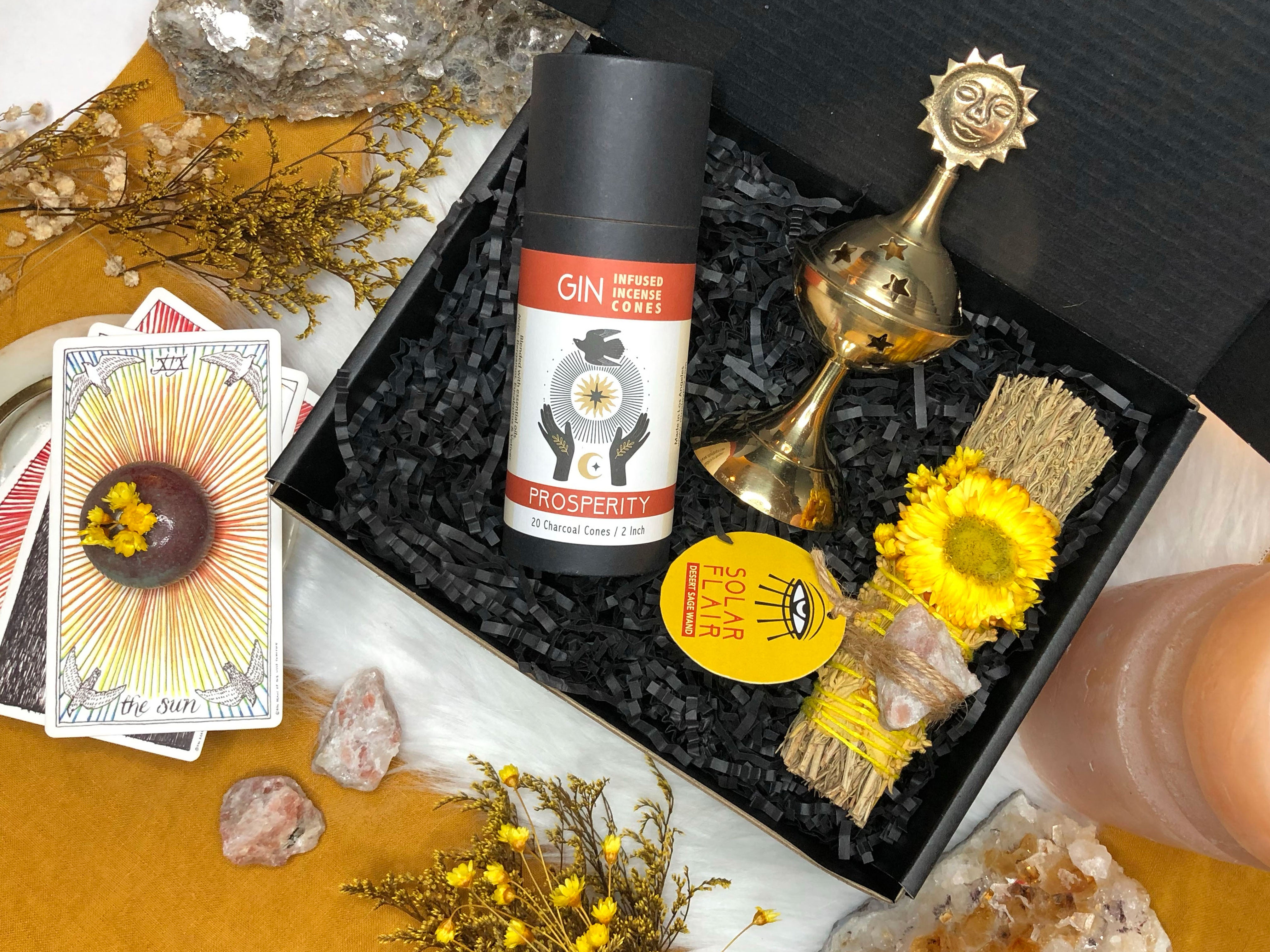 black git box that has brass sun incense holder, desert sage smoke cleansing bundle with yellow rose petals and yellow straw flower and sunstone attached and black container with gin infused incense cones and tarot cards