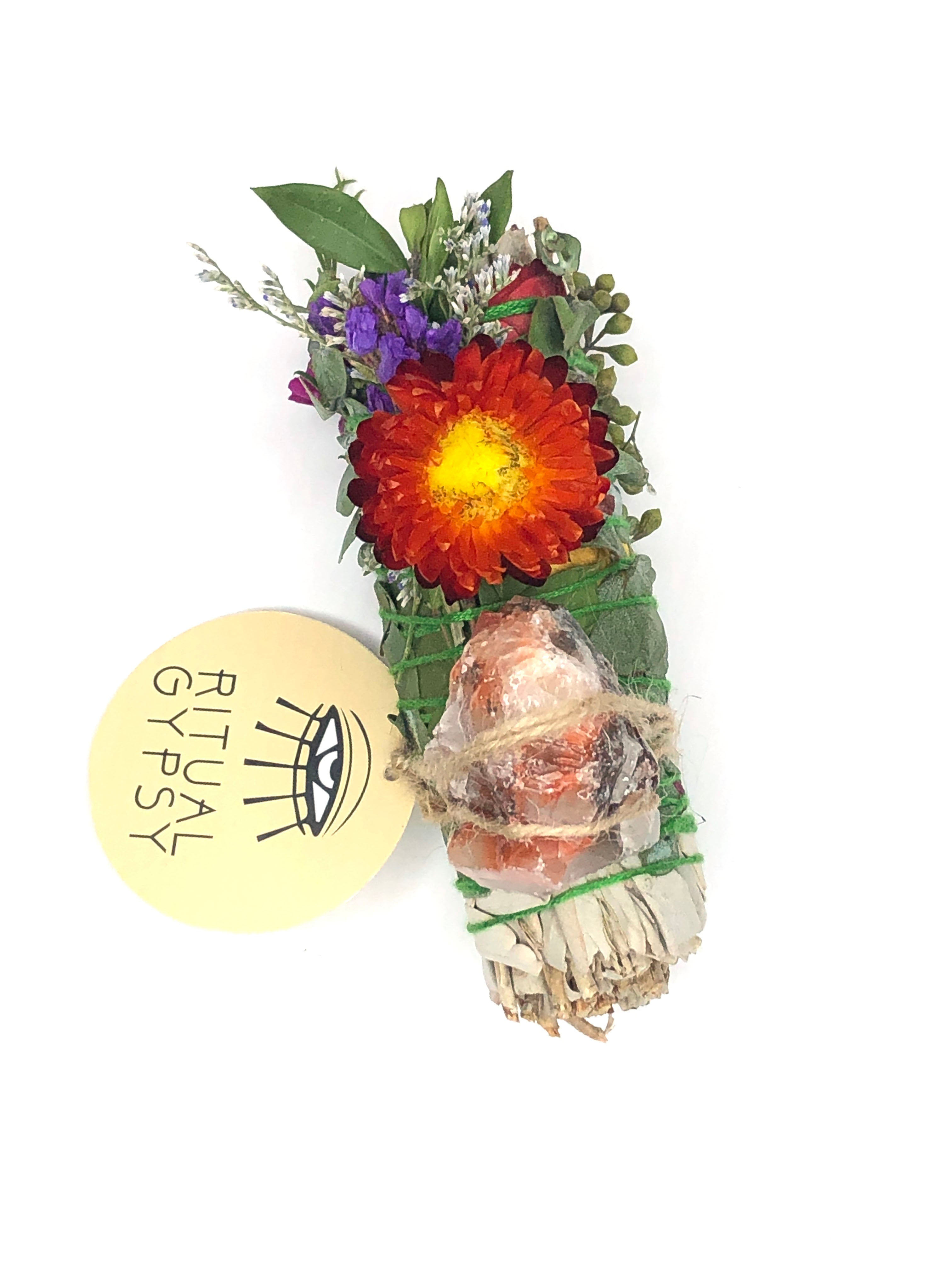 white sage smoke cleansing smudge bundle wrapped in florals with red strawflower and orange rainbow calcite crystal and round ritual gypsy product tag attached on white background